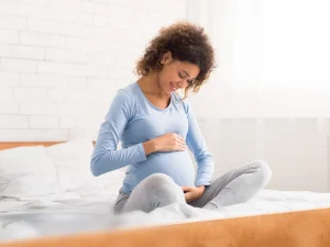 Pregnant woman sitting on bed, smiling and holding her belly